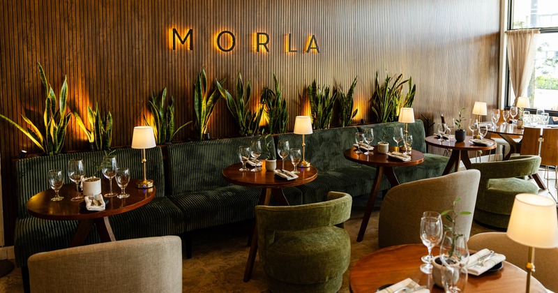 A modern restaurant interior with wooden paneling, green seating, round tables, and ambient lighting