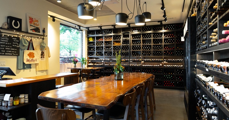 Warmly lit  interior - a long wooden table, chairs, merchandise displays, walls with wine bottles