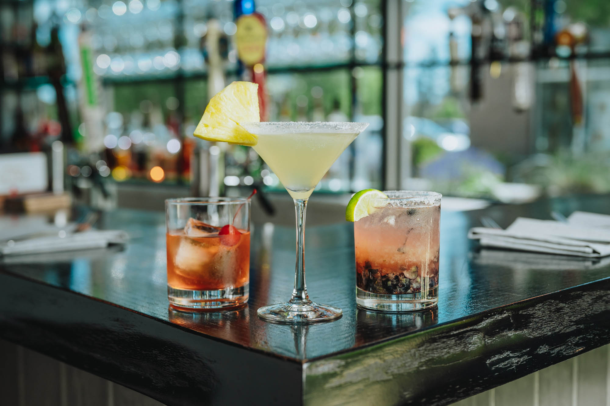 Three colorful cocktails with fruit garnishes sitting on a bar counter