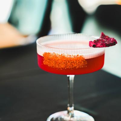 Red cocktail with white foam, orange-rim seasoning and dried fruit garnish in a clear ribbed glass.