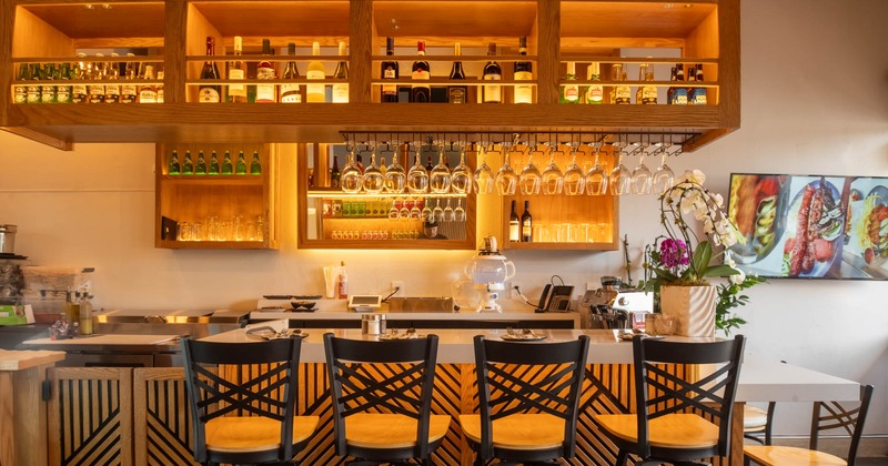 A warm-lit bar area with hanging wine glasses, liquor bottles on shelves, and high chairs