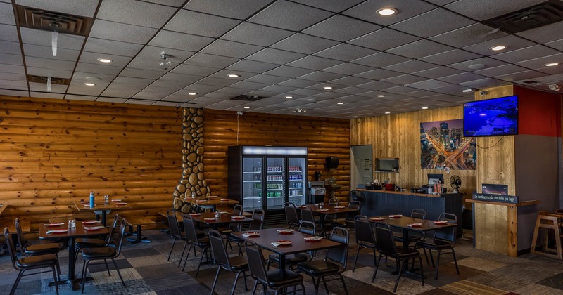 Wood-paneled restaurant interior with empty tables and chairs, a small counter, fridges, and a TV