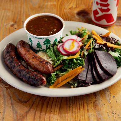 A plate with two sausages, a side green salad with beets, and a cup of baked beans.