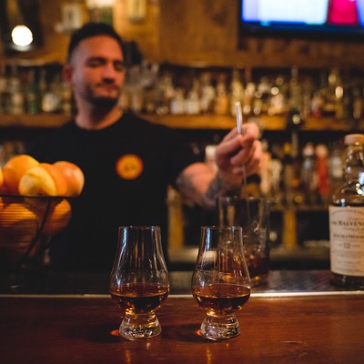 Two Glencairn glasses with straight whiskey on the bar with a bartender in the background.