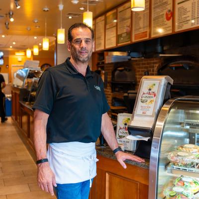 A person in a black shirt and white apron by the pizzeria counter.