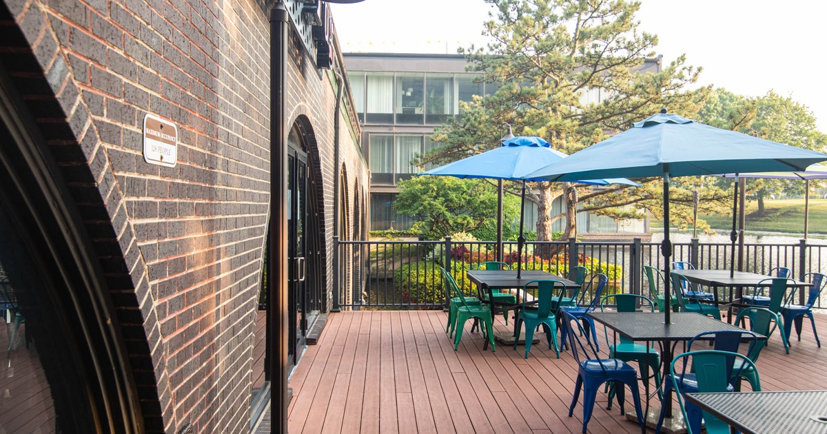 Outdoor patio with tables, chairs, and umbrellas next to a brick building and a body of water