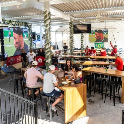 Outdoor dining area with large TV screens amd people seated at high-top tables.