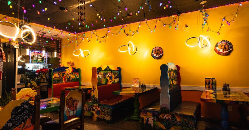 A restaurant interior with yellow walls, colorful murals on booth backrests, and multicolored string lights overhead