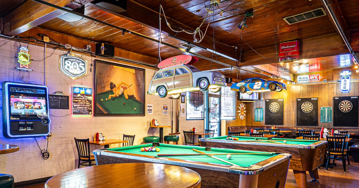 Interior of a bar with two pool tables, neon signs and a painting of a woman at a pool table.