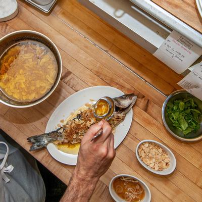 Whole sea bass, hand pouring a dressing over the dish, side dishes, top view