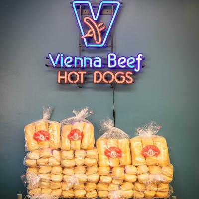 A neon 'Vienna Beef Hot Dogs' sign above stacks of packaged buns.