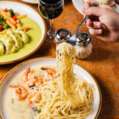 A hand twirls spaghetti on a shrimp scampi plate, assorted food plates and red wine.