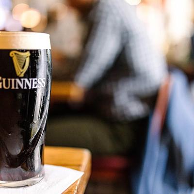 A glass of Guinness sits on a wooden table, with blurred patrons in the background.