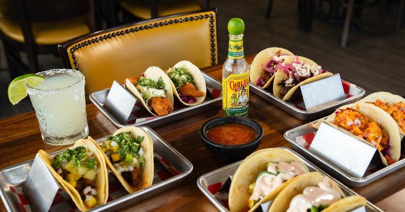 A spread of assorted tacos served in metal holders on metal trays alongside drinks