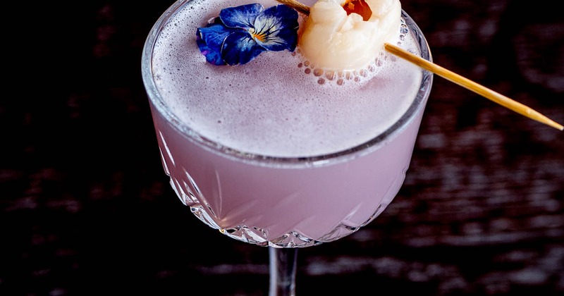 A frothy purple cocktail in a crystal glass, garnished with a blue flower and lychee on a skewer