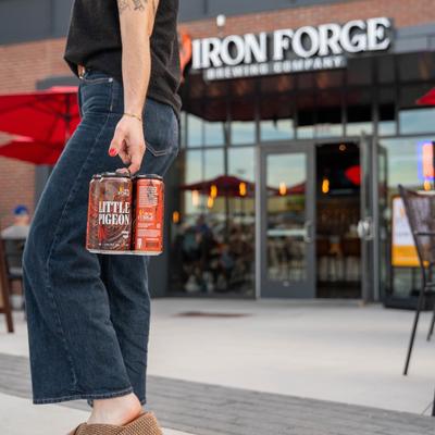 Person holding a six-pack of beer with a storefront and signage in the background.