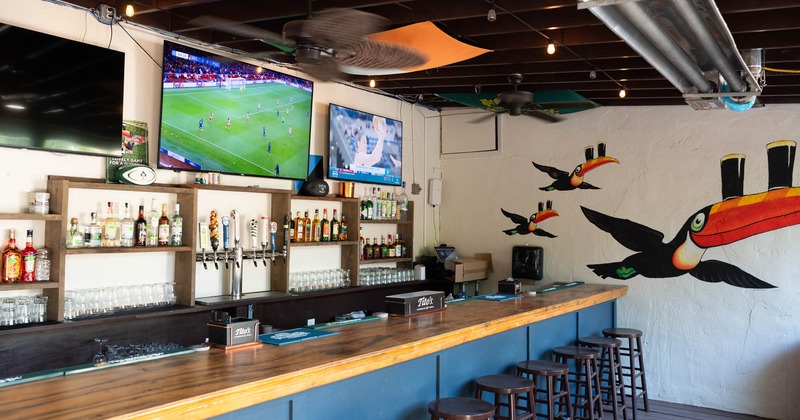 Bar interior with a long counter, shelves with bottles, TVs displaying sports, ceiling flags