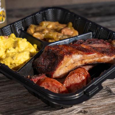 BBQ rib with sausage, potato salad, and smothered green beans in a takeout tray.