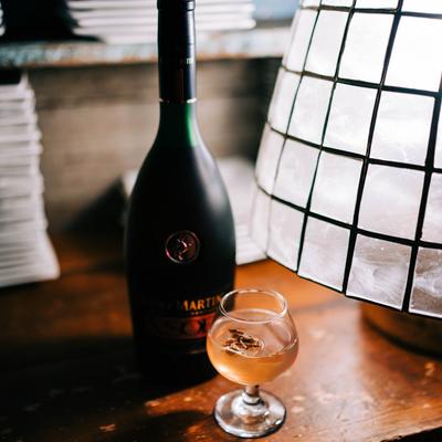 A bottle of cognac and a full glass on a table next to a lamp.