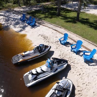 Three jet skis on a sandy beach beside blue chairs and trees.