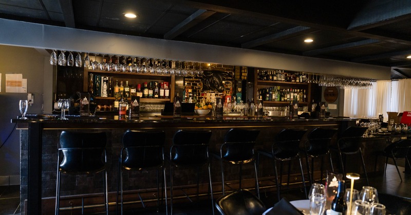 Dark upscale bar stocked with liquor bottles and hanging wine glasses, with black bar stools.