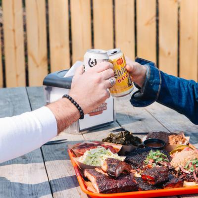 Outdoors, two people toasting with beer at a table with smoked meat platter.