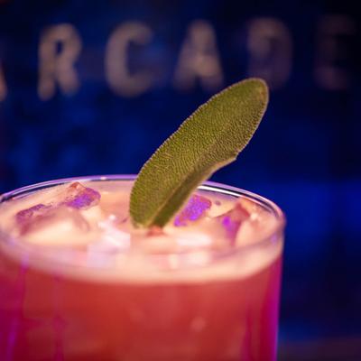 Close-up of a pink cocktail with a fresh sage leaf garnish, under a soft purple light.