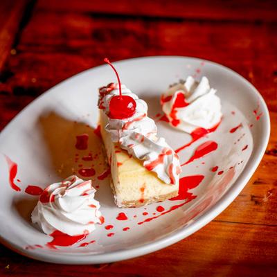 A slice of cheesecake with whipped cream, cherry, and red syrup drizzle on a plate.