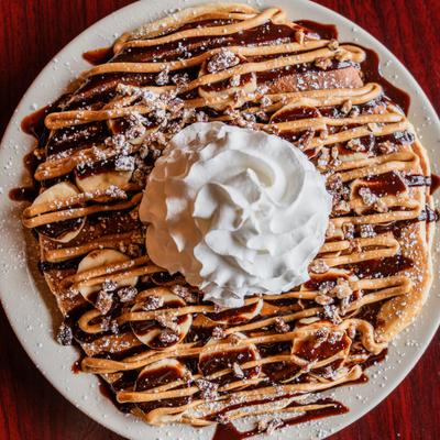 Smores Pancakes topped with banana slices, chocolate chips, sauces and whipped cream.