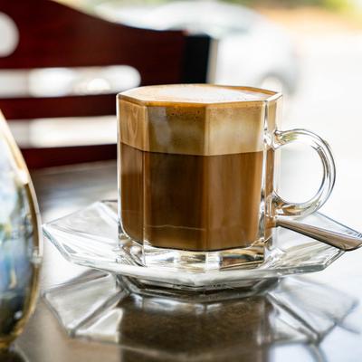 A glass mug with frothy macchiato  sits on a reflective table inside.