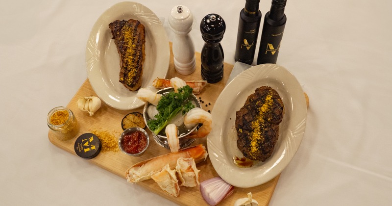 Grilled steaks sit on a wooden board with  shrimp cocktail, crab legs, spices, and condiments