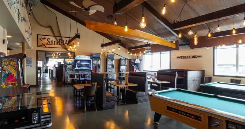 Interior with booths, gaming machines, a pool table, and hanging light bulbs, with natural light from large windows