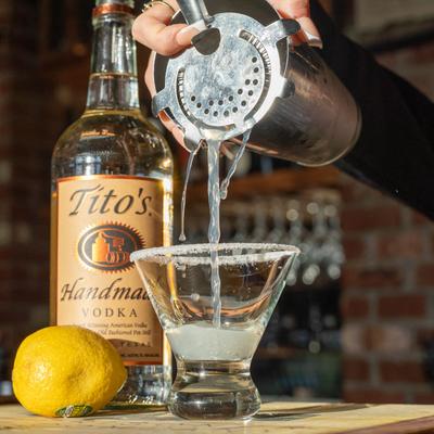 Bartender straining a cocktail into a glass, lemon and a bottle of vodka nearby.