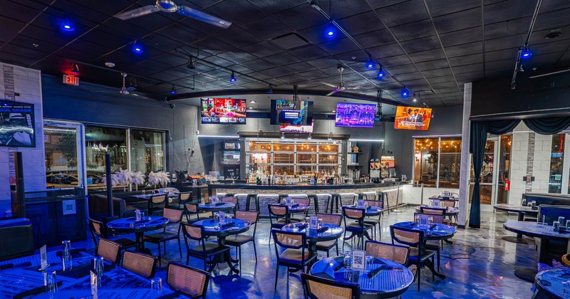 Modern restaurant interior with round tables, black chairs, and a bar with multiple TVs