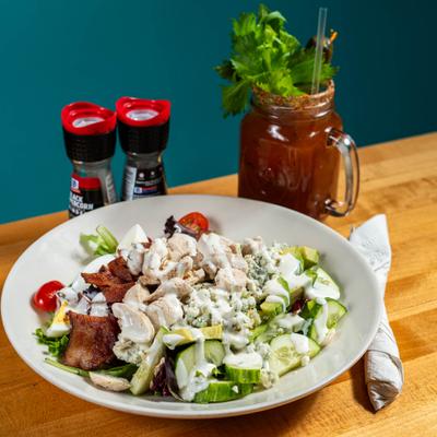 Large salad with chicken, bacon, and vegetables, set on a table with condiments and cocktail.