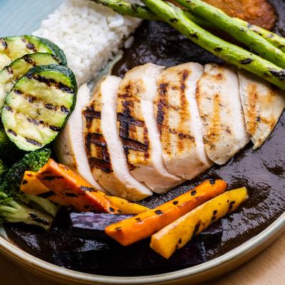 Close-up of grilled chicken with vegetables and rice over dark sauce.