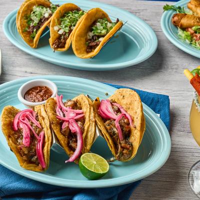 Steak tacos with pink onions, served with lime, salsa, and street tacos in the background.