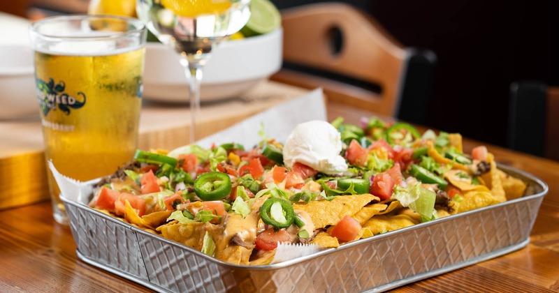 A metal tray with nachos, accompanied with a glass of beer and a cocktail