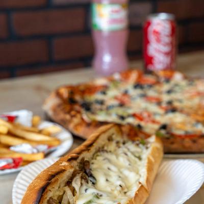 Philly cheesesteak sub sandwich with fries, pizza, and sodas in the background.