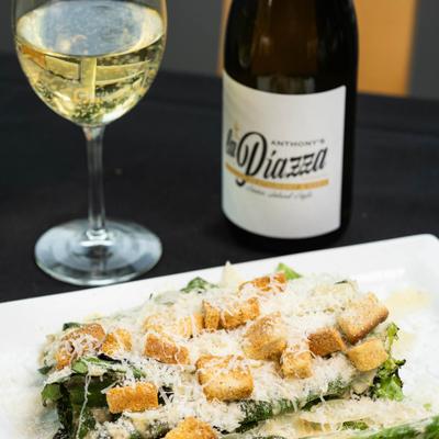 A plate holds Caesar Salad on a dark table with a glass and bottle of white wine.