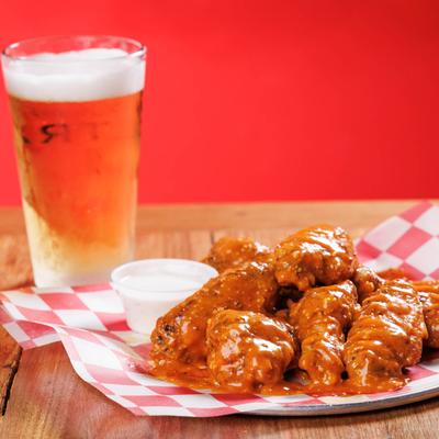 A plate of saucy wings on red-checkered paper sits on a wooden table beside a glass of beer.