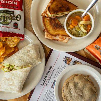Lunch spread with a grilled sandwich, chicken wrap, soup, potato chips, and a cookie.