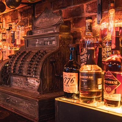 Assorted bottles of liquor lined up at a bar, with an old cash register in view.