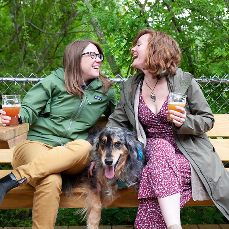 Two people sitting on a bench outdoors with a dog, holding drinks and smiling.