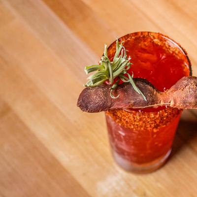 Spicy red cocktail with chili rim and bacon garnish.