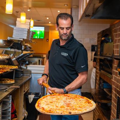 A pizza maker holds a large cheese pizza on a peel in a pizzeria kitchen.