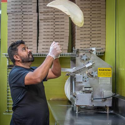 A person tossing pizza dough with stacks of pizza boxes in the background.