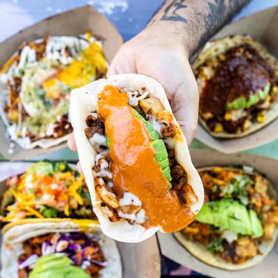 Hand holding a taco topped with orange sauce, above the table with assortment of tacos.