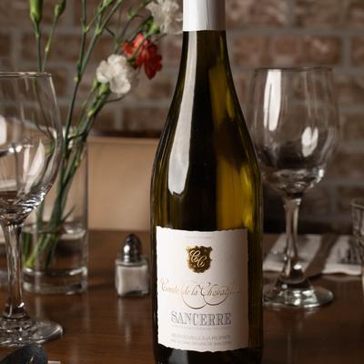 A bottle of white wine sits on a wooden table with a bouquet of carnations and wine glasses.