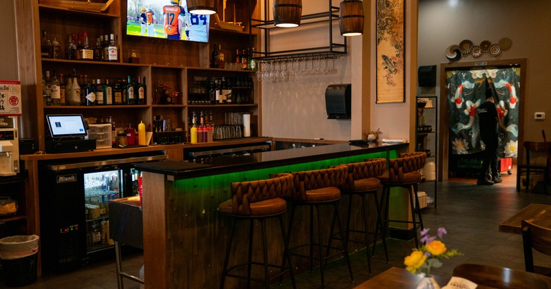 Warmly lit bar - liquor shelves, pendant lights, green-lit counter, bar stools, TV